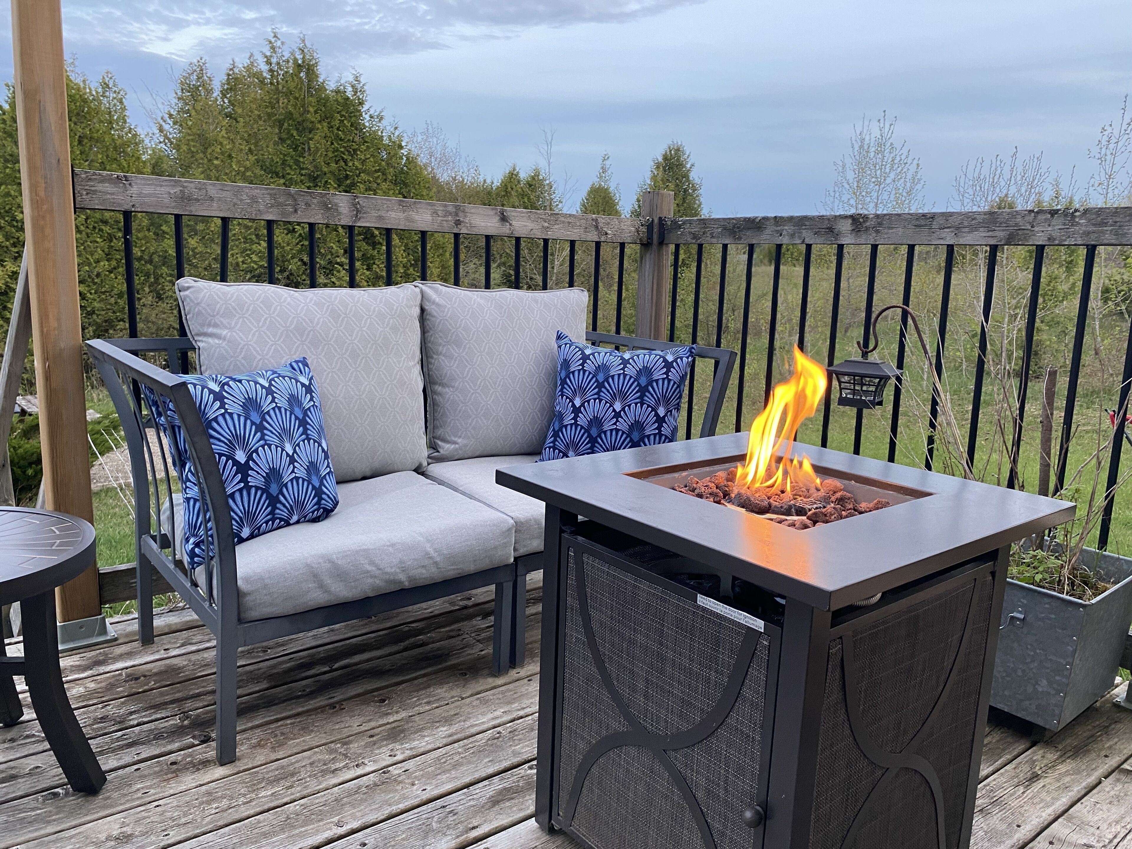 A deck with a gray sofa, blue patterned pillows, and a square fire pit with flames.