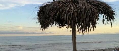 A beach with a thatched umbrella and two lounge chairs.