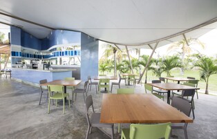 Uma área de refeições ao ar livre com mesas e cadeiras, um bar e palmeiras ao fundo.