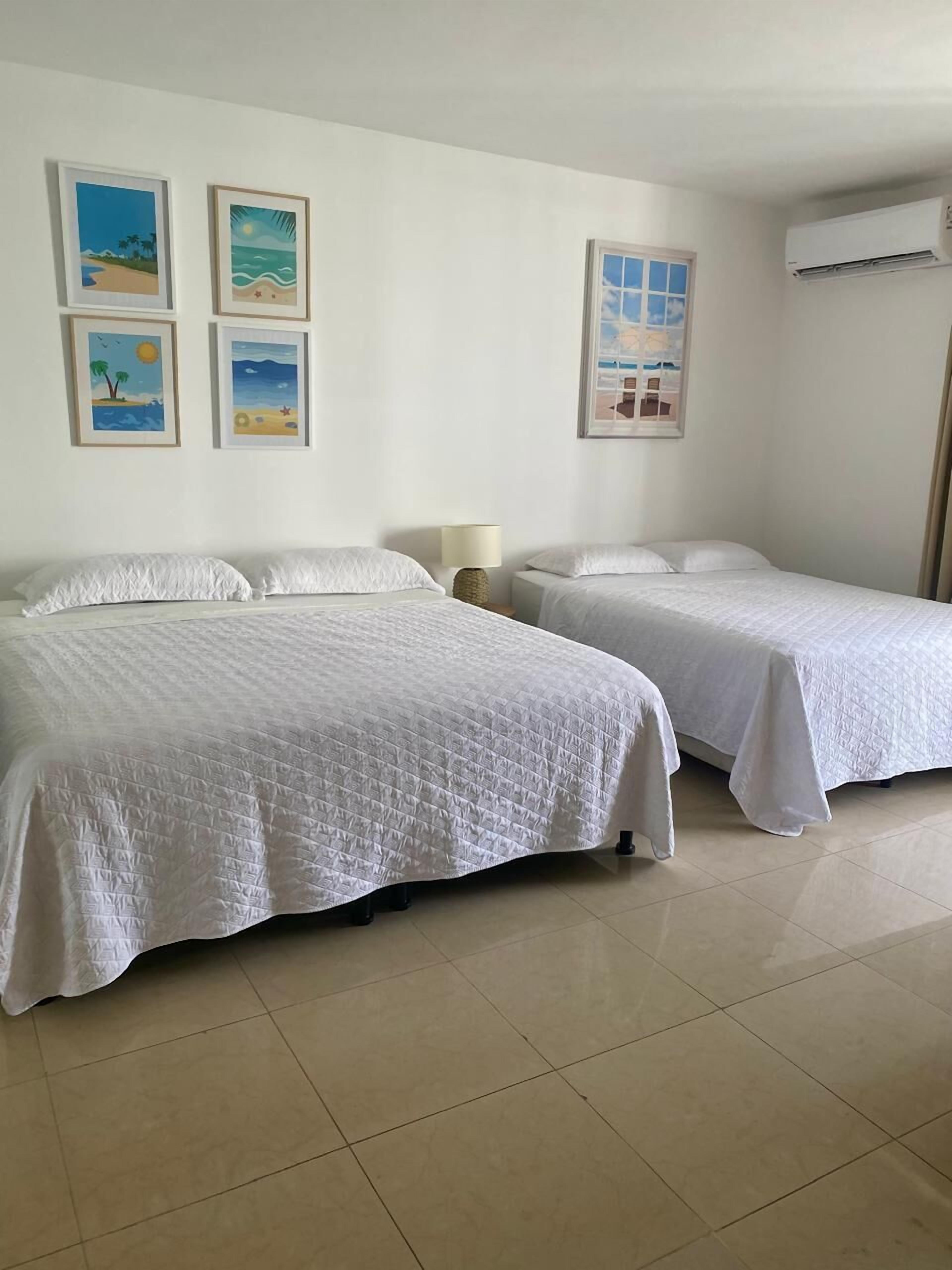 Una habitación de hotel con dos camas, una mesita de noche, una lámpara y cuatro cuadros en la pared.