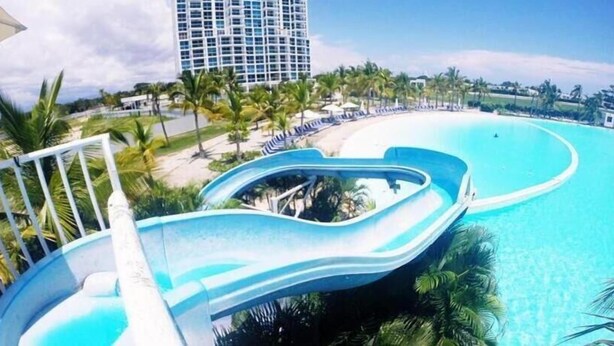 A swimming pool with a water slide, palm trees, and a large building in the background. - Apartamento en Playa Blanca Town Center (Rio Hato)
