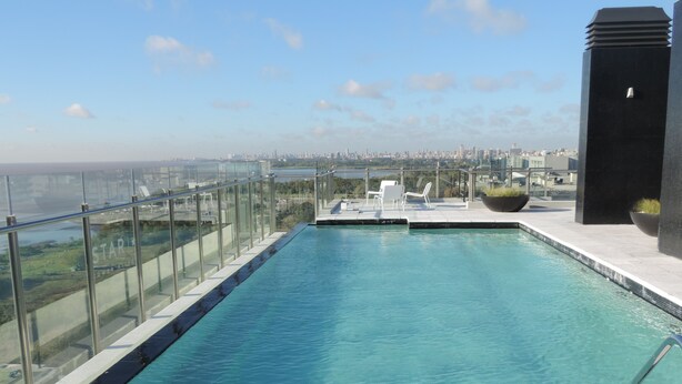 Una piscina infinita en la azotea con agua azul cristalina, rodeada por barandillas de vidrio, sillas de descanso blancas y vistas al horizonte de la ciudad.