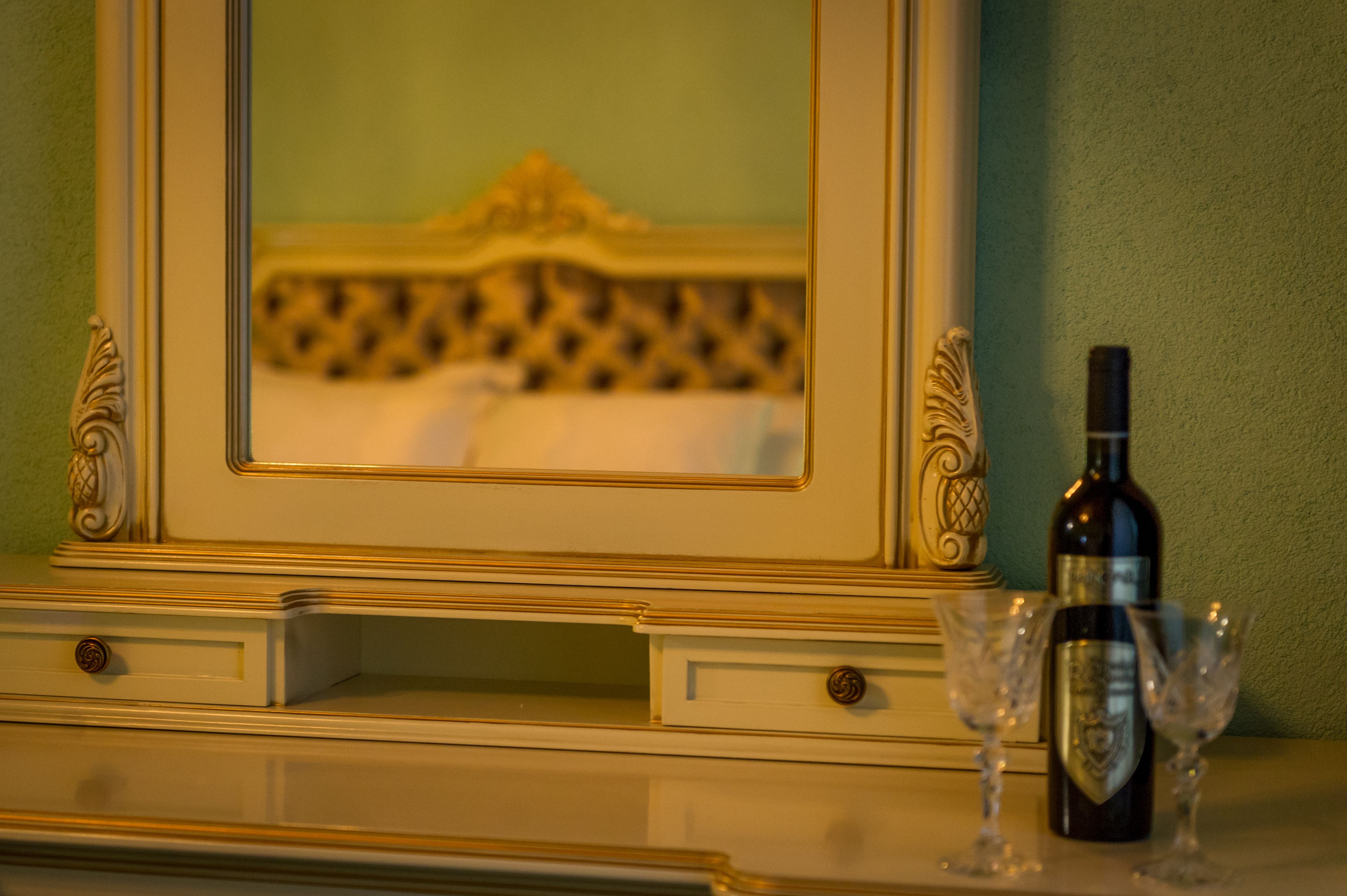 A mirror with a gold frame, a bottle of wine, and two wine glasses on a dresser.