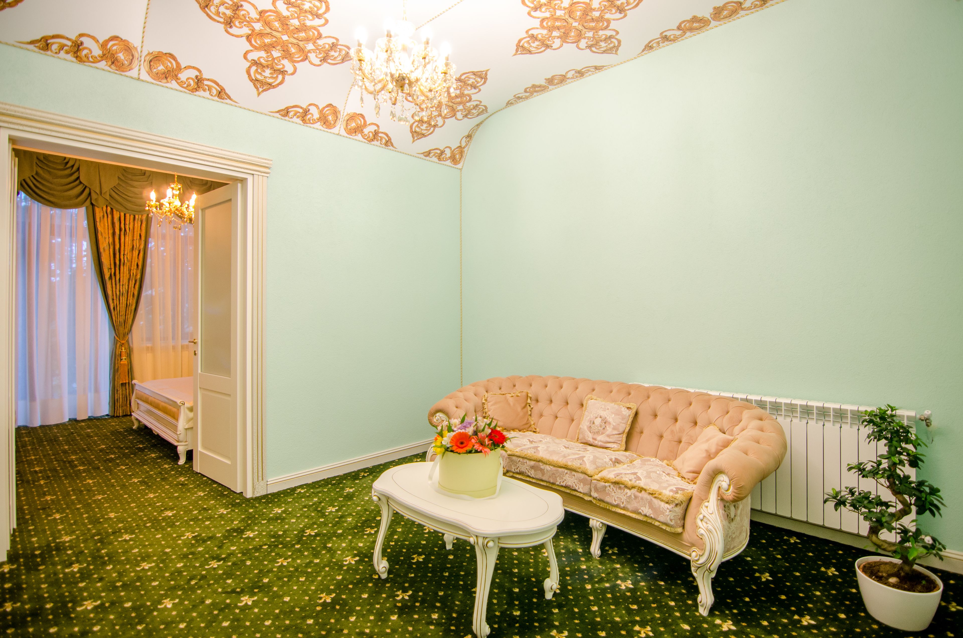 A room with a green carpet, a white sofa, a small table with a vase of flowers, a radiator, and a potted plant.