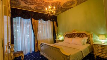 A hotel room with a large bed, a nightstand with a lamp, a bench, and a view of the outdoors through the window.