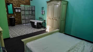 A hotel room with a bed, a small table, a wardrobe, and a bathroom with a patterned shower.
