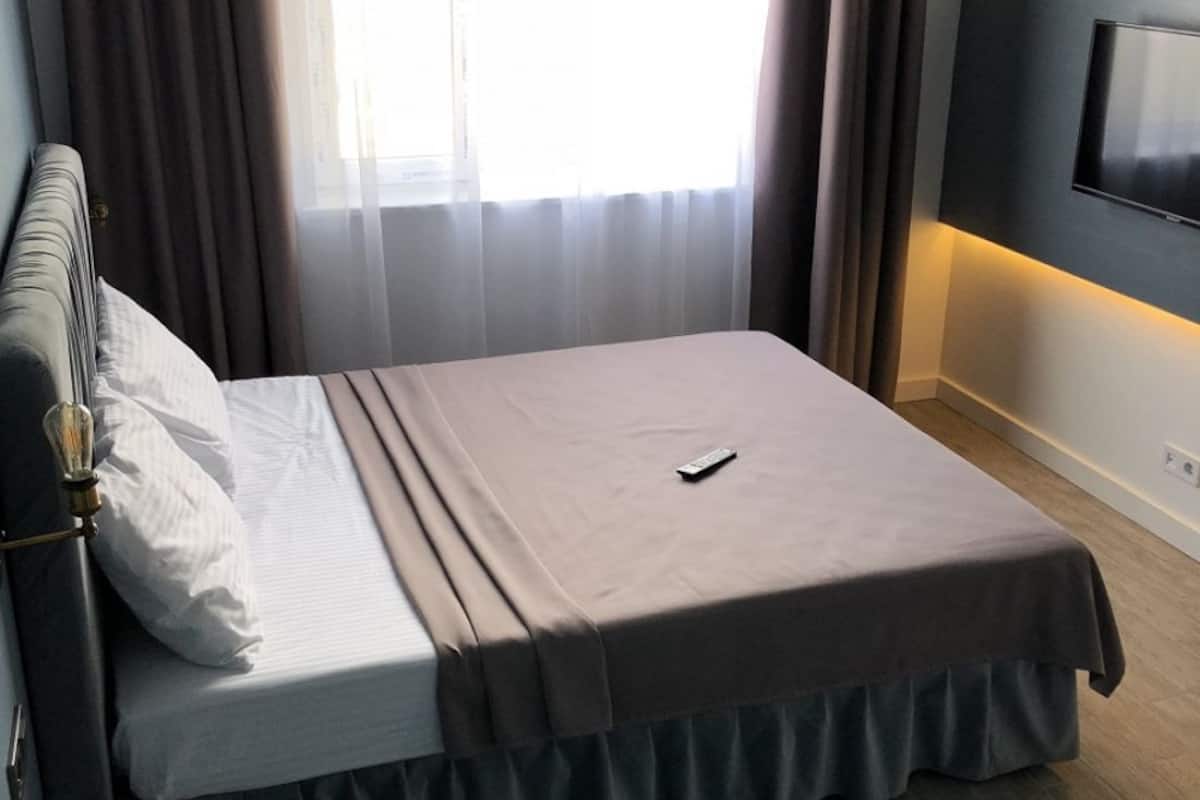 A neatly made bed with a remote control on it, a TV mounted on the wall, and a window with sheer curtains.