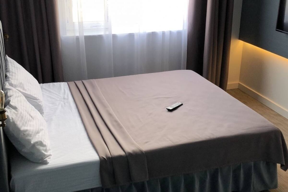 A neatly made bed with a remote control on it, a window with sheer curtains, and a wall-mounted light fixture.