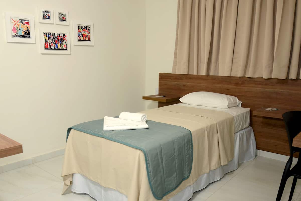 Un lit bien fait, recouvert d’une courtepointe beige et turquoise, avec une tête de lit en bois et un oreiller blanc. On peut voir des tableaux encadrés au mur, une table de chevet en bois et une chaise.