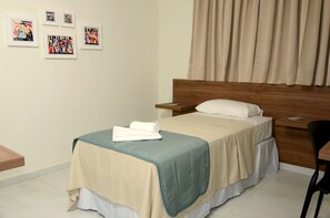 A neatly made bed with a beige and teal bedspread, a wooden headboard, and a white pillow. There are framed pictures on the wall, a wooden nightstand, and a chair.