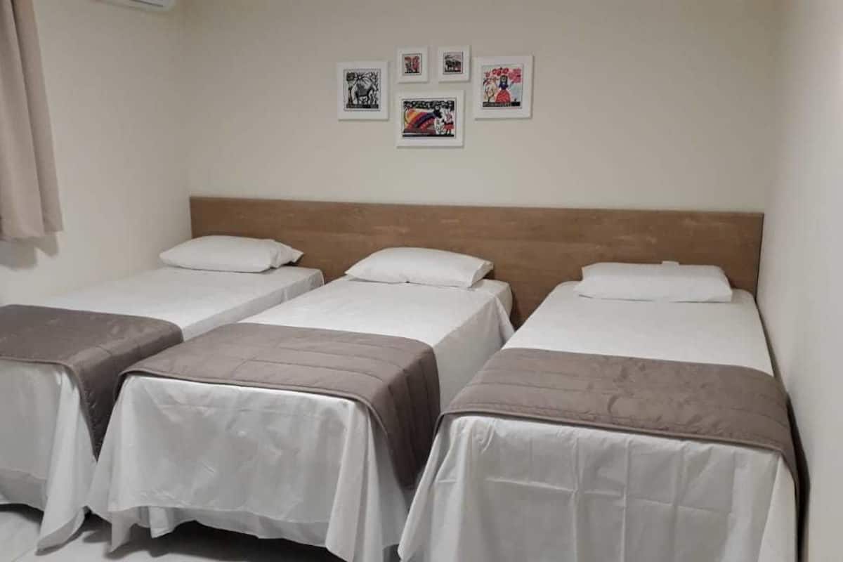 Une chambre avec trois lits simples, chacun doté d’un matelas blanc et d’une tête de lit marron. Quatre tableaux encadrés sont accrochés au mur, au-dessus des lits.