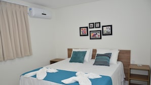 Una habitación de hotel con una cama, una colcha azul, almohadas blancas y una manta azul. Hay cuadros en la pared y un equipo de aire acondicionado sobre la cama.