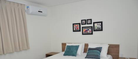 A hotel room with a bed, a blue bedspread, white pillows, and a blue blanket. There are framed pictures on the wall and an air conditioning unit above the bed.