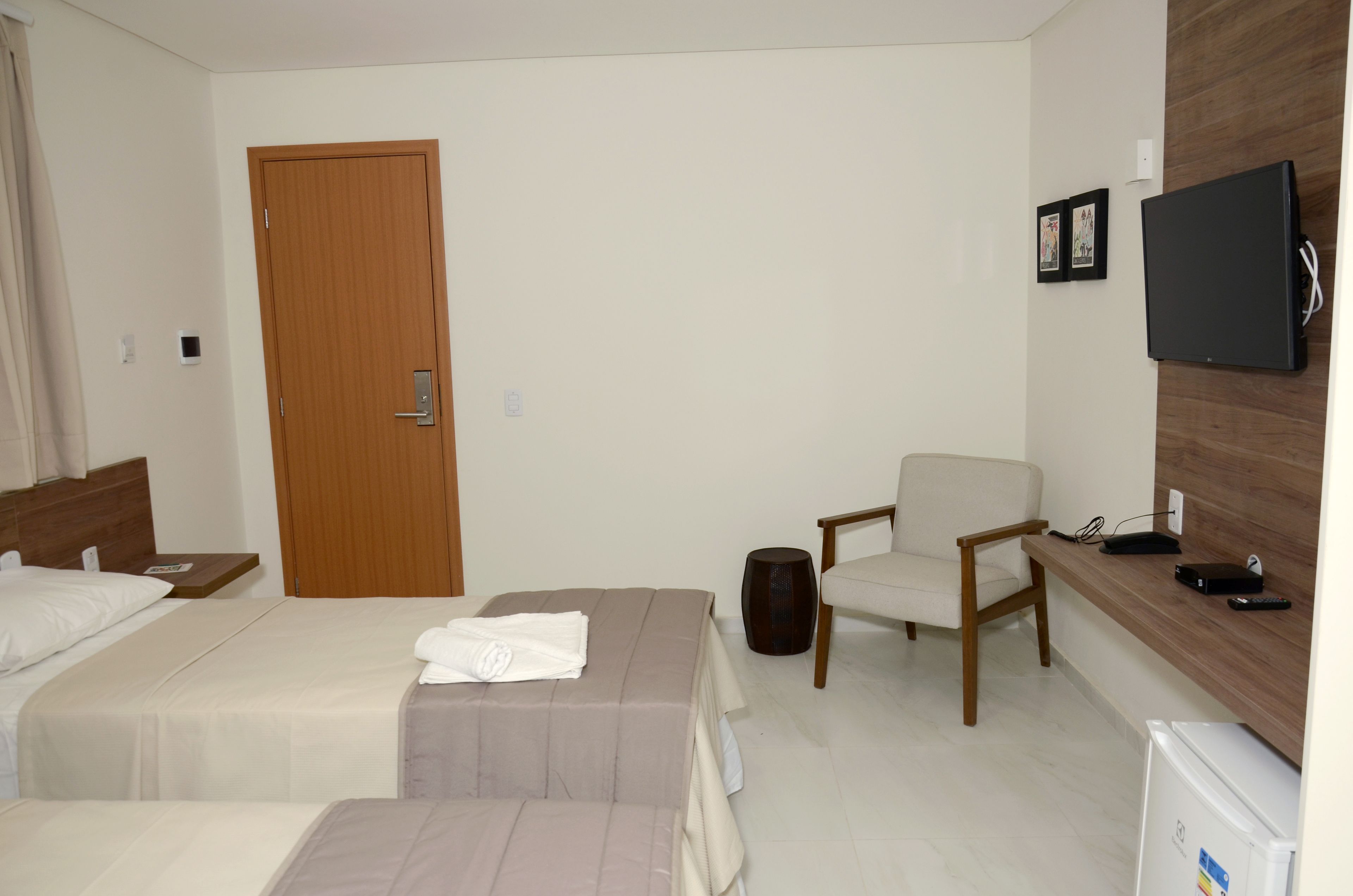 Quarto de hotel com duas camas, uma porta de madeira, televisão fixada na parede, uma cadeira e uma mesa com computador e telefone.