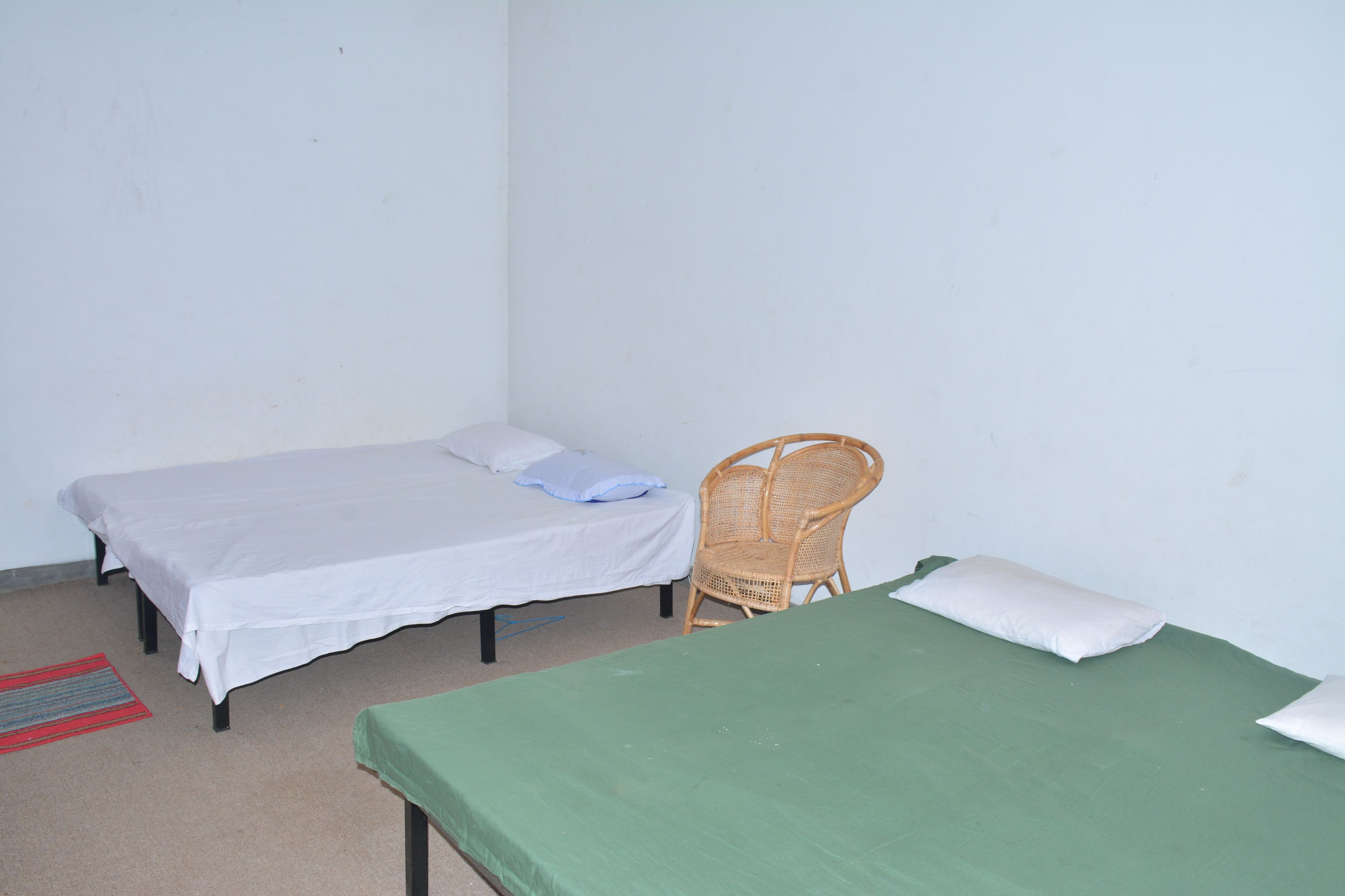Two single beds with white and green bedding, a wicker chair, and a red and gray striped rug in a simple room with plain walls.