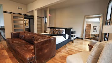 A hotel room with a large bed, a brown leather sofa, a small round table, and a wooden floor.