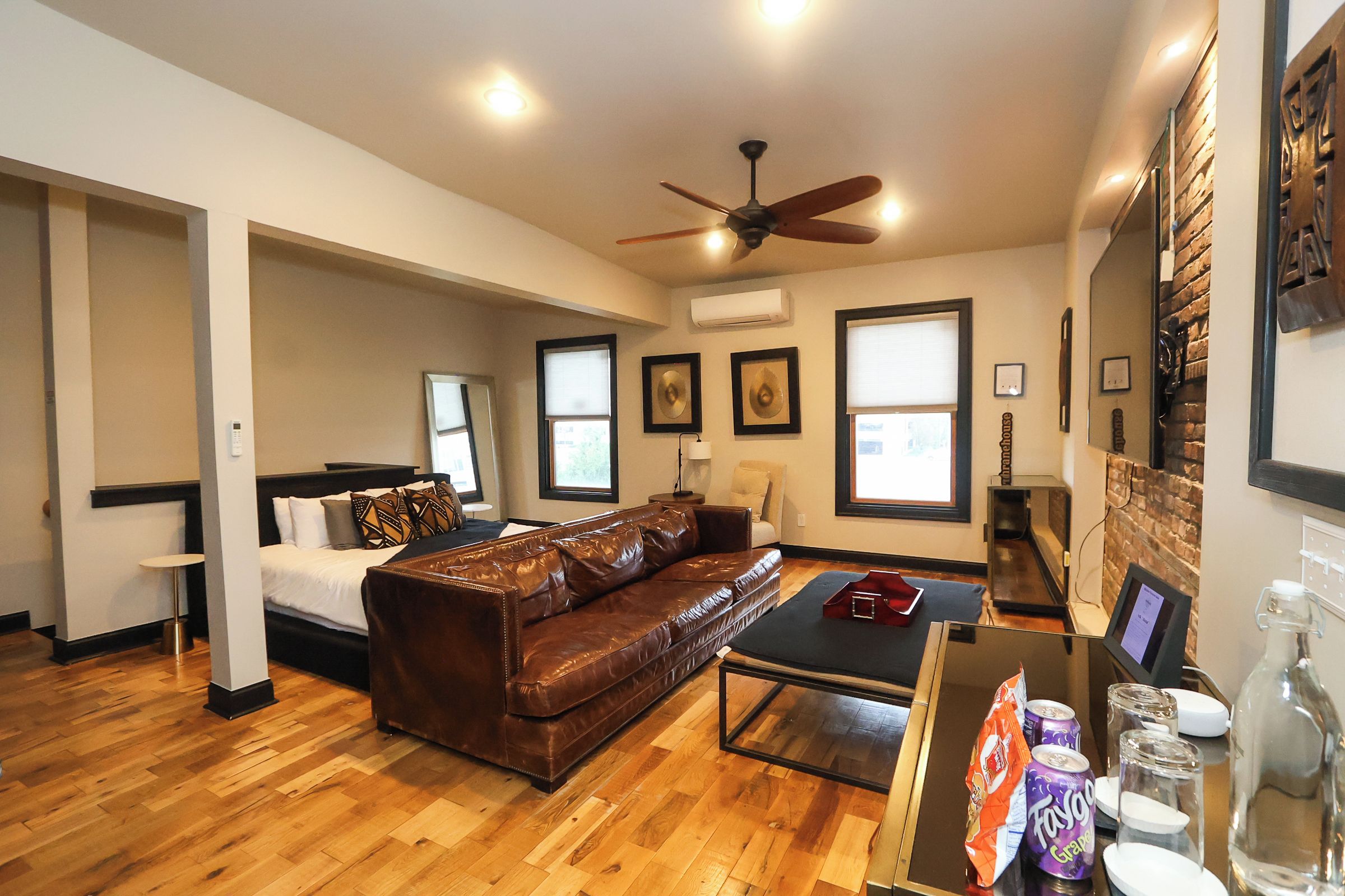 A spacious living room with a brown leather sofa, a coffee table, and a ceiling fan. There are windows with blinds, a fireplace, and a wall-mounted TV.
