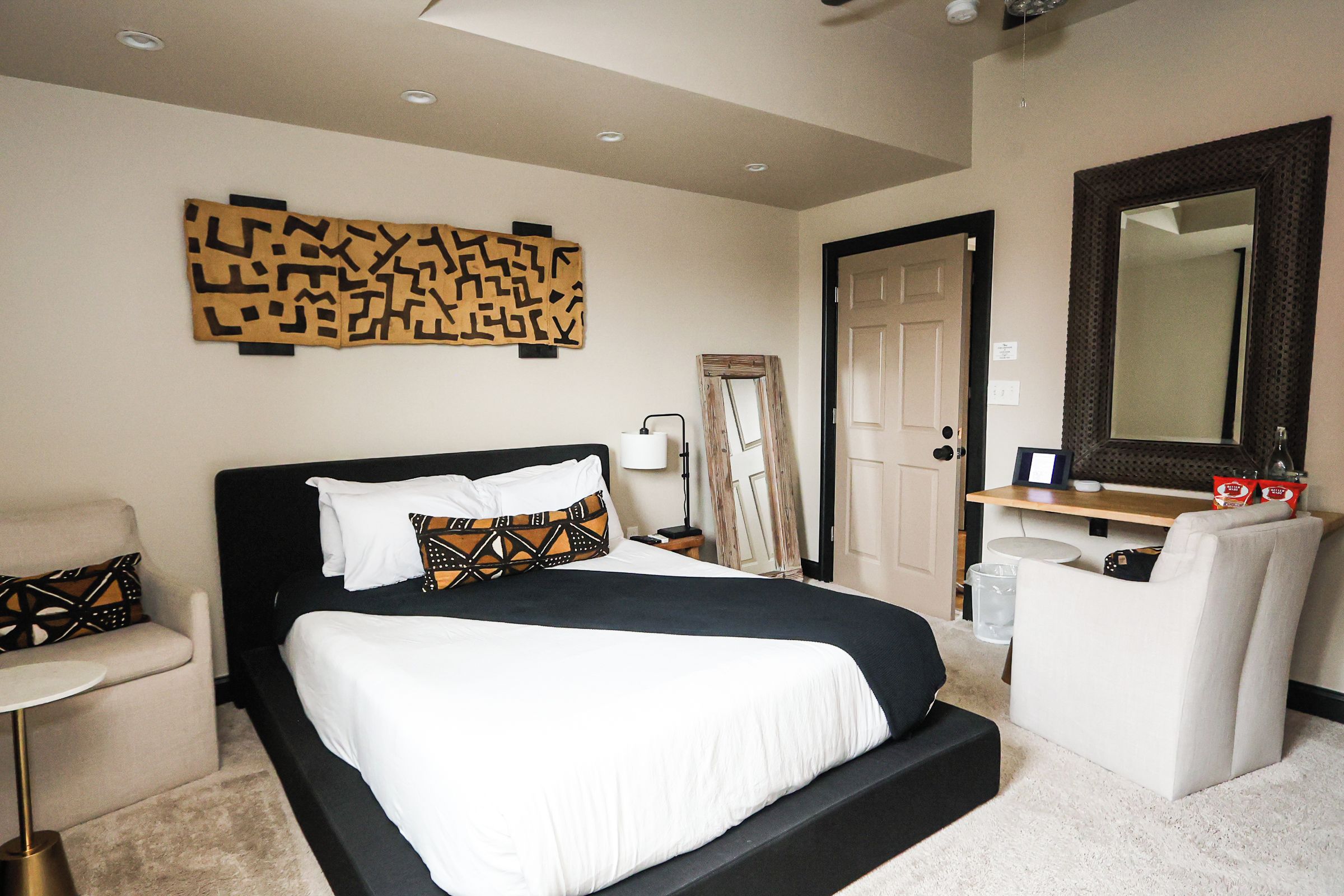 A hotel room with a large bed, a chair, a desk, a mirror, and a wall art piece.