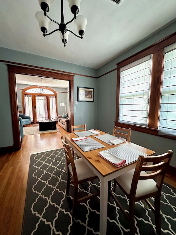 A dining area with a wooden table, chairs, and a patterned rug. There is a chandelier above the table and a window with blinds on the right. - Mid City 2 BR in Beautiful Arts and Crafts House (New Orleans)