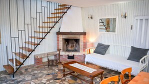 Una habitación acogedora con suelo de piedra, una escalera de madera, chimenea, sofá blanco y una mesa de comedor.