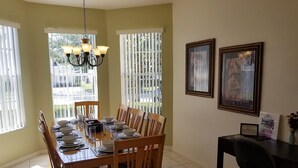 A dining area with a wooden table set for four, surrounded by chairs. There is a chandelier above the table, and the room has large windows with blinds. Two framed pictures are on the wall to the right. - Stay in Luxury, Private Pool/Spa, Games Room, Close to Disney and Golf (Haines City)