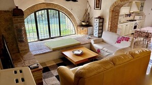 A living room with a sofa, a coffee table, a fireplace, a kitchen area, and a large arched window. - Esclusivo per te (COLLE VAL D' ELSA)