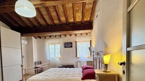 A bedroom with a bed, a bedside table with a lamp, a chair, a window with curtains, and a wooden ceiling. - Esclusivo per te (COLLE VAL D' ELSA)