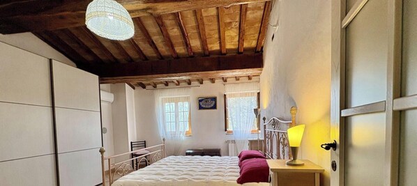 Ein Schlafzimmer mit einem Bett, einem Nachttisch mit Lampe, einem Sessel, einem Fenster mit Vorhängen und einer Holzbalkendecke.