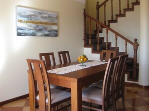 A dining area with a wooden table, six chairs, and a glass bowl of fruit on the table. A staircase with wooden railings is visible in the background. - Sunny Villa - Sea View, BBQ & Private Pool (Ponta do Sol)