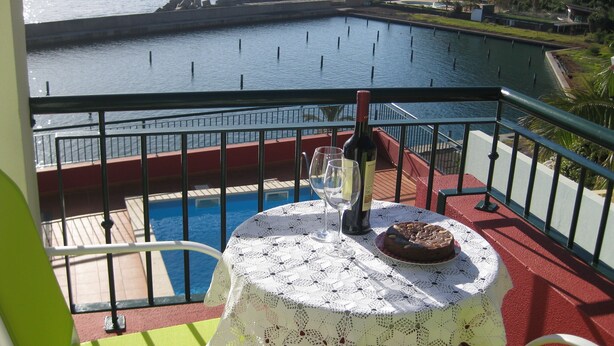 Un balcón con una mesa preparada para dos personas, una botella de vino y un pastel. El balcón tiene vista a un cuerpo de agua y una costa rocosa.