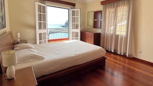 Un dormitorio con una cama grande, una mesita de noche, un aparador y vistas al océano a través de una ventana abierta.