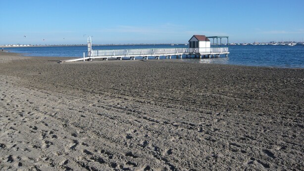 Una spiaggia con un molo che si protende nell'acqua, un piccolo edificio bianco all'estremità e un cielo azzurro terso.