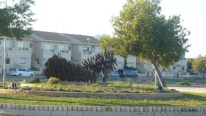 Una scultura in un’area verde con una strada e edifici residenziali sullo sfondo.