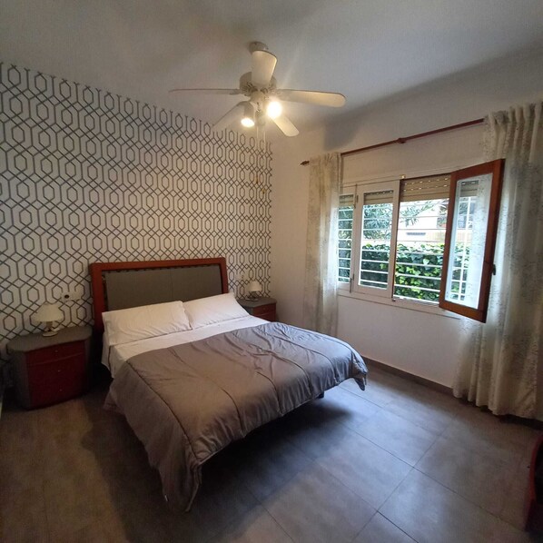 A bedroom with a bed, a ceiling fan, a window with curtains, and a patterned wall.