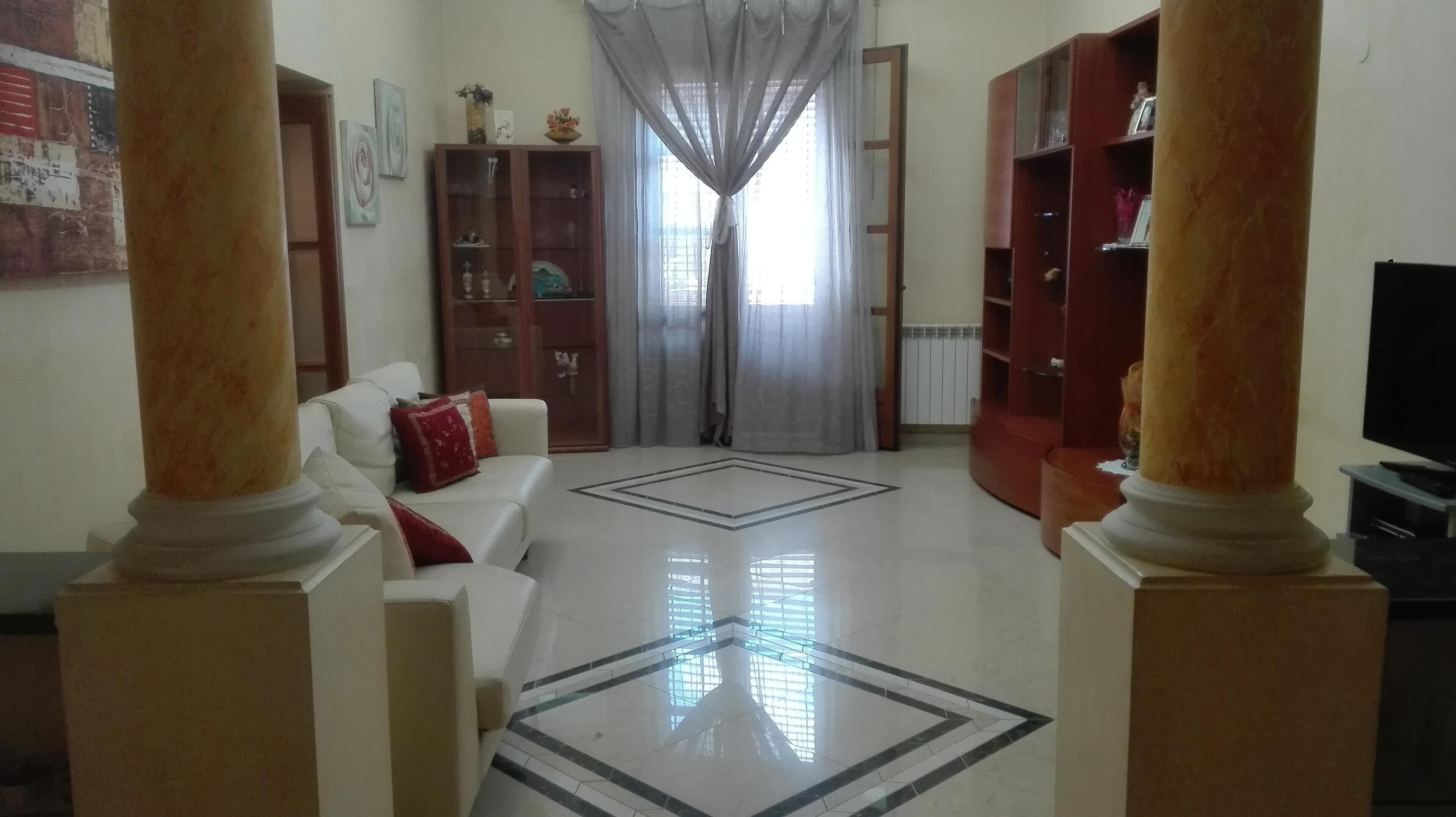 Un salón con un sofá blanco, cojines rojos, una mesa de centro de vidrio y un televisor. Hay dos estanterías de madera y un ventanal con cortinas traslúcidas.