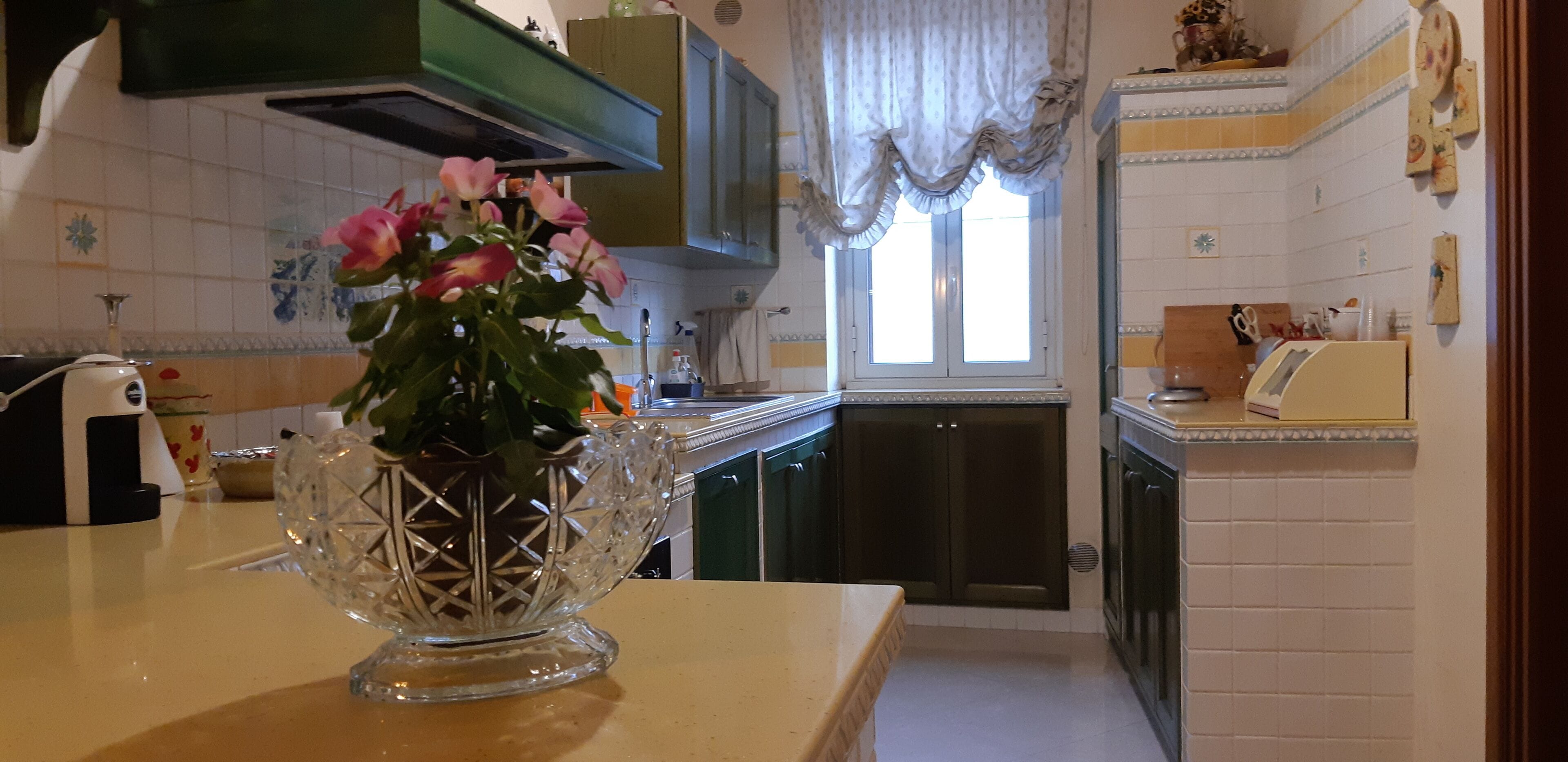 Une cuisine avec des meubles de couleur verte, une fenêtre avec des rideaux et un vase rempli de fleurs posé sur le comptoir.