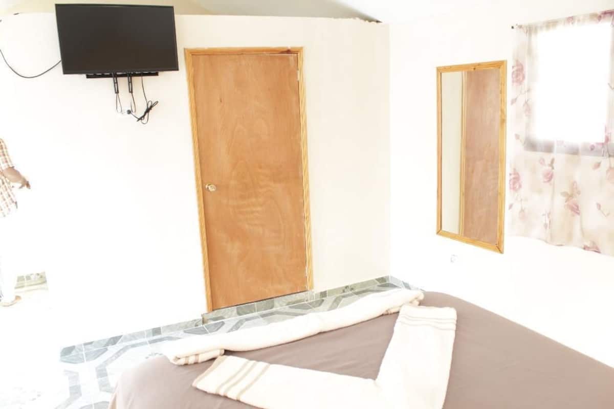 Une pièce comprenant un lit, une porte en bois, un téléviseur fixé au mur et une fenêtre ornée de rideaux à motifs floraux.