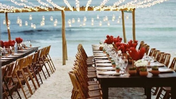 An outdoor dining setup with string lights overhead, wooden chairs, and tables arranged on a sandy beach with the ocean in the background. - Evasion (Gressier)