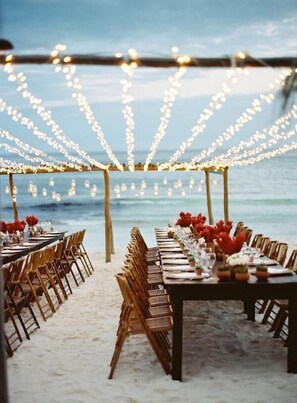An outdoor dining setup with string lights overhead, wooden chairs, and tables arranged on a sandy beach with the ocean in the background. - Evasion (Gressier)