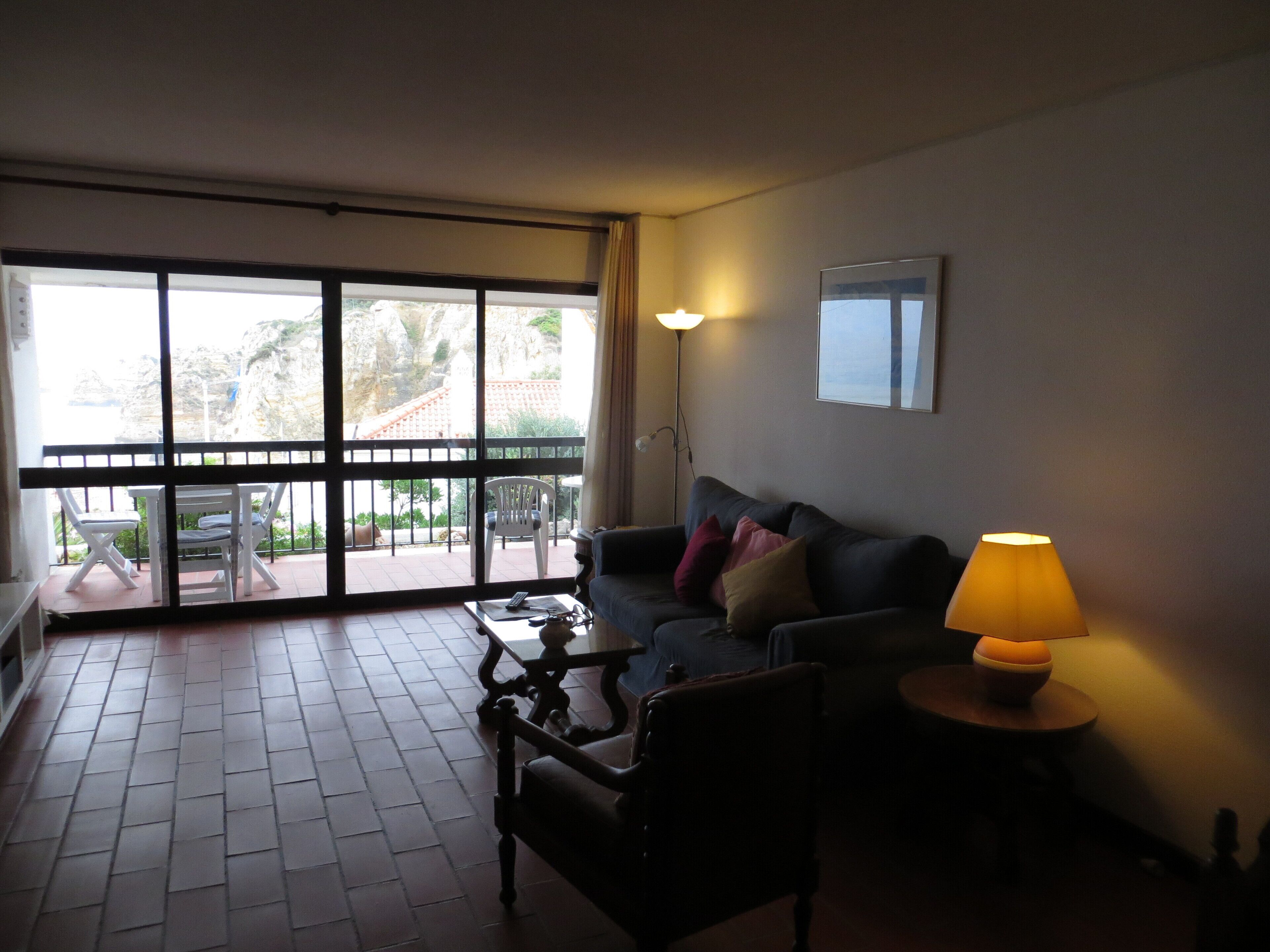 A living room with a sofa, a coffee table, and a lamp. There is a balcony with chairs and a table outside.