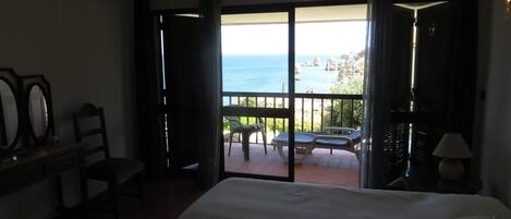 A hotel room with a bed, a desk, a chair, and a balcony with a view of the sea.