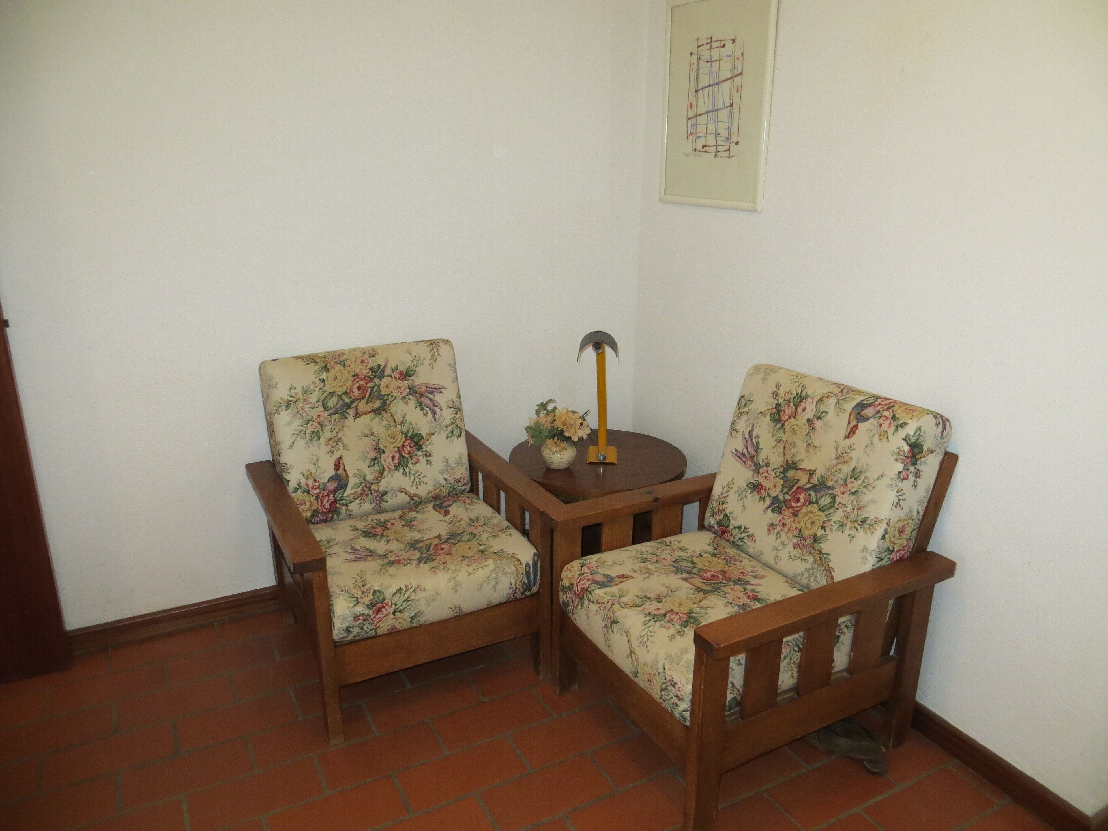Two wooden chairs with floral upholstery, a small wooden table with a vase, and a framed abstract artwork on the wall.
