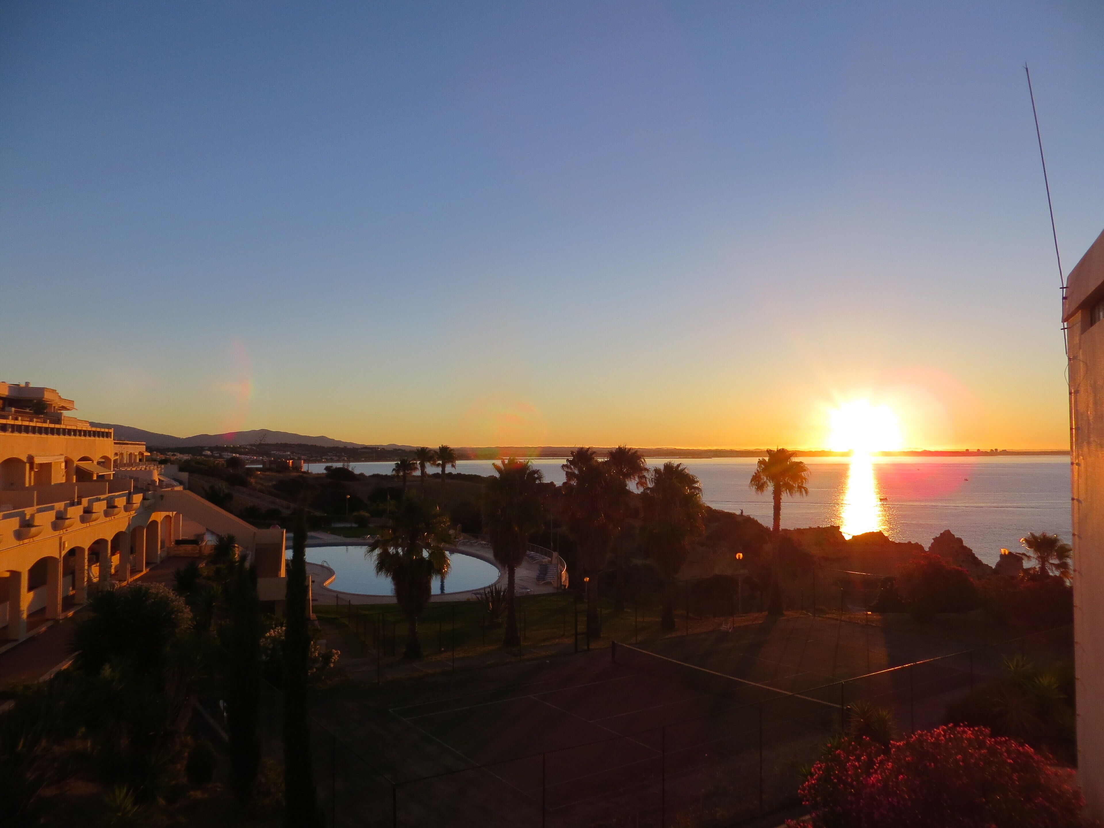 A sunset view from a hotel balcony overlooking a pool and palm trees.