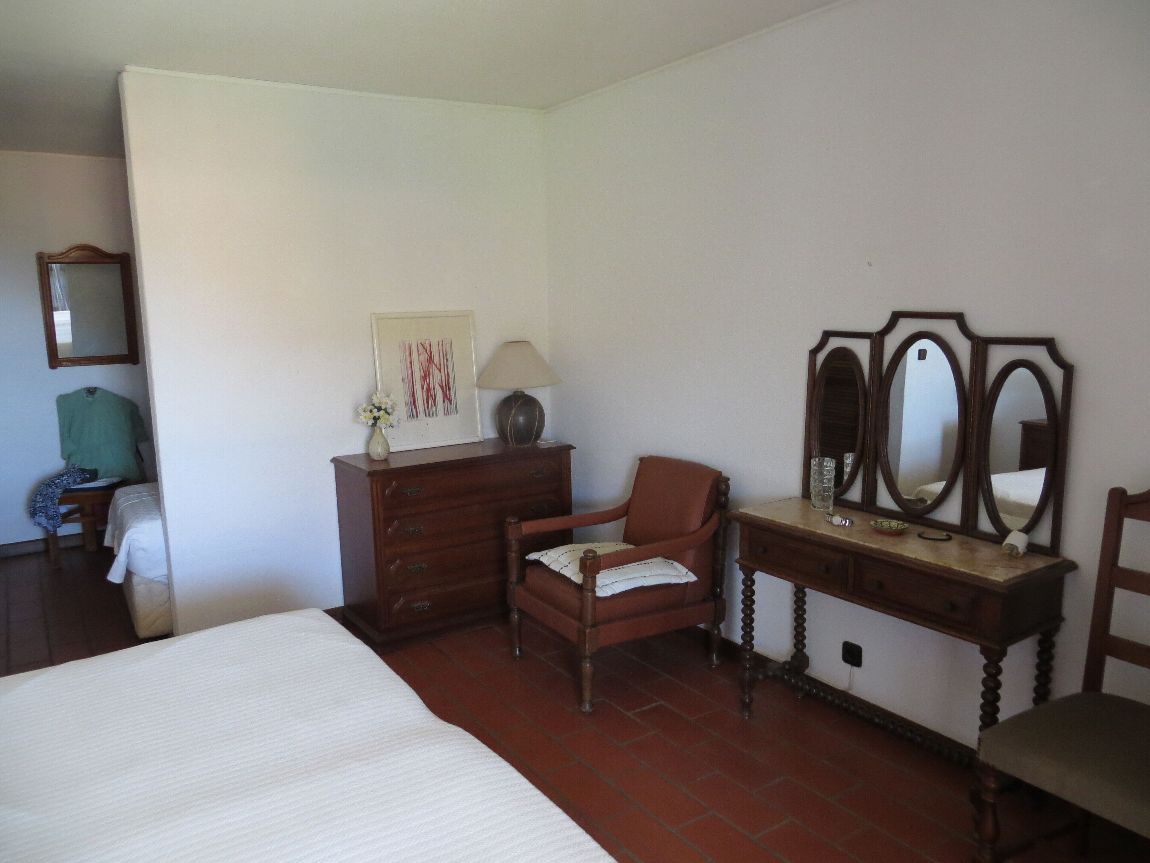 A bedroom with a bed, a wooden dresser, a chair, a desk with a mirror, and a framed picture on the wall.