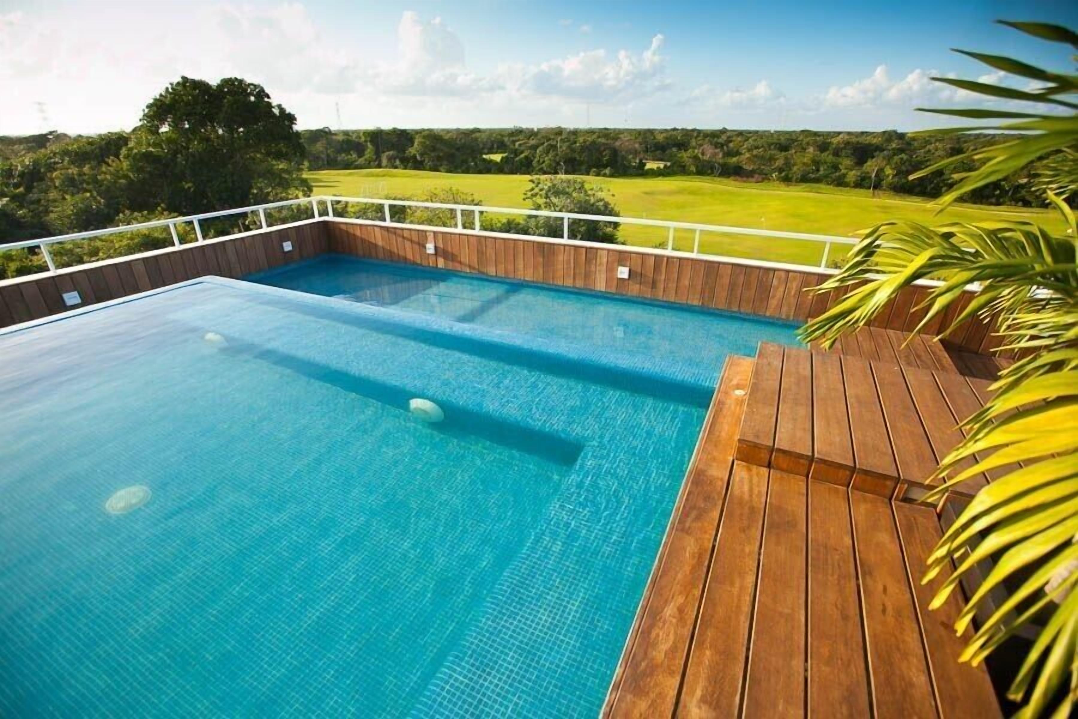 Ein rechteckiger Pool mit Holzterrasse und weißer Reling, umgeben von üppiger Vegetation und blauem Himmel.