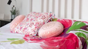 A neatly made bed with floral bedding and decorative pillows, a nightstand with a lamp and a plant, and a painting of two figures on a beach above the bed. - Modern Apartment "Woodleigh" Sleeps 4 (London)