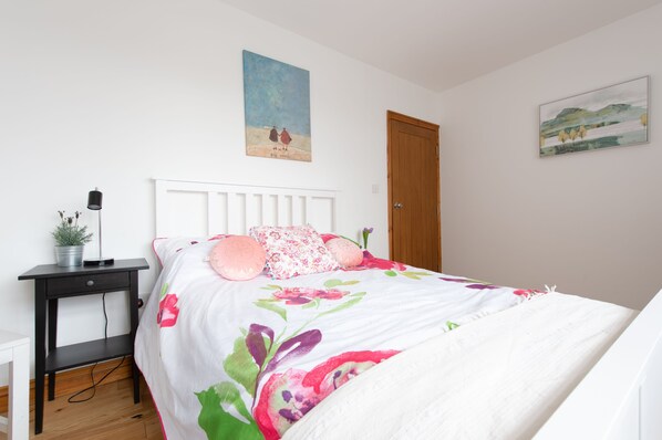 Quarto com cama branca, roupa de cama floral, mesa de cabeceira com abajur e duas pinturas na parede.