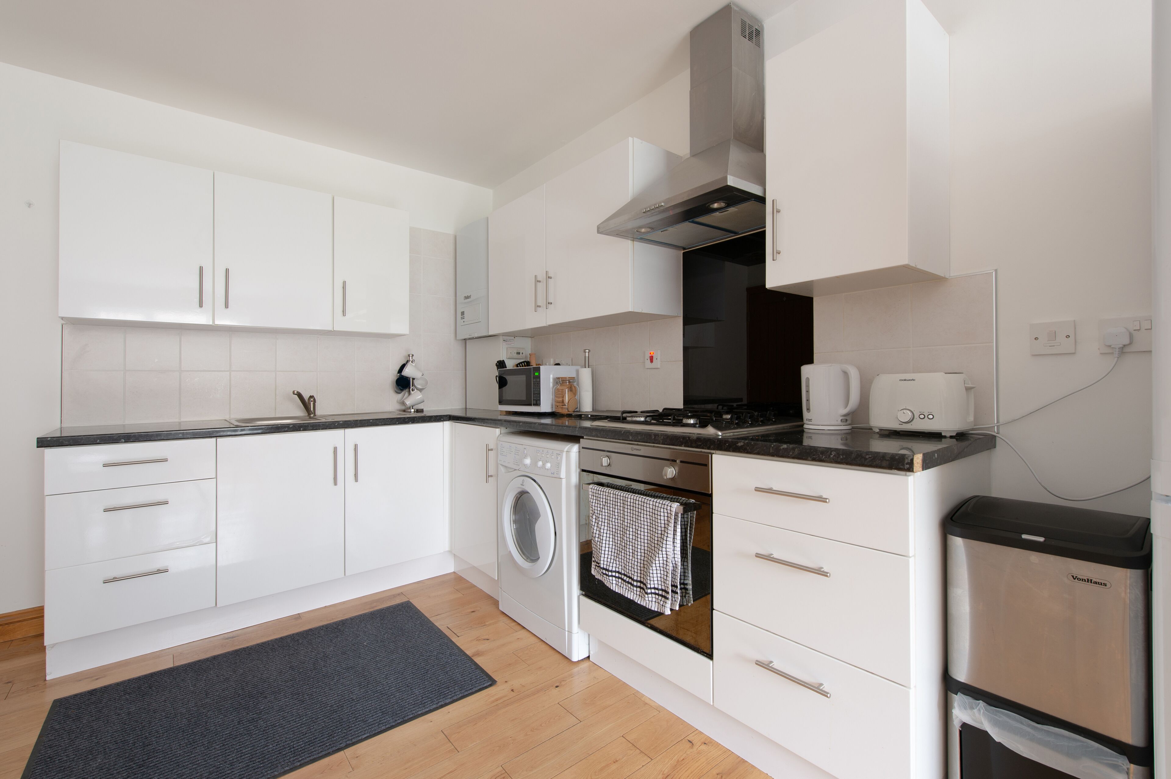 Una cocina moderna con muebles blancos, lavadora integrada, estufa y cafetera.