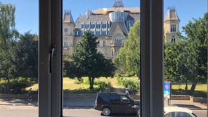 A view through a window of a building with a large, ornate structure in the background, a paved area with trees, and a black car parked on the street. - Modern Apartment "Woodleigh" Sleeps 4 (London)