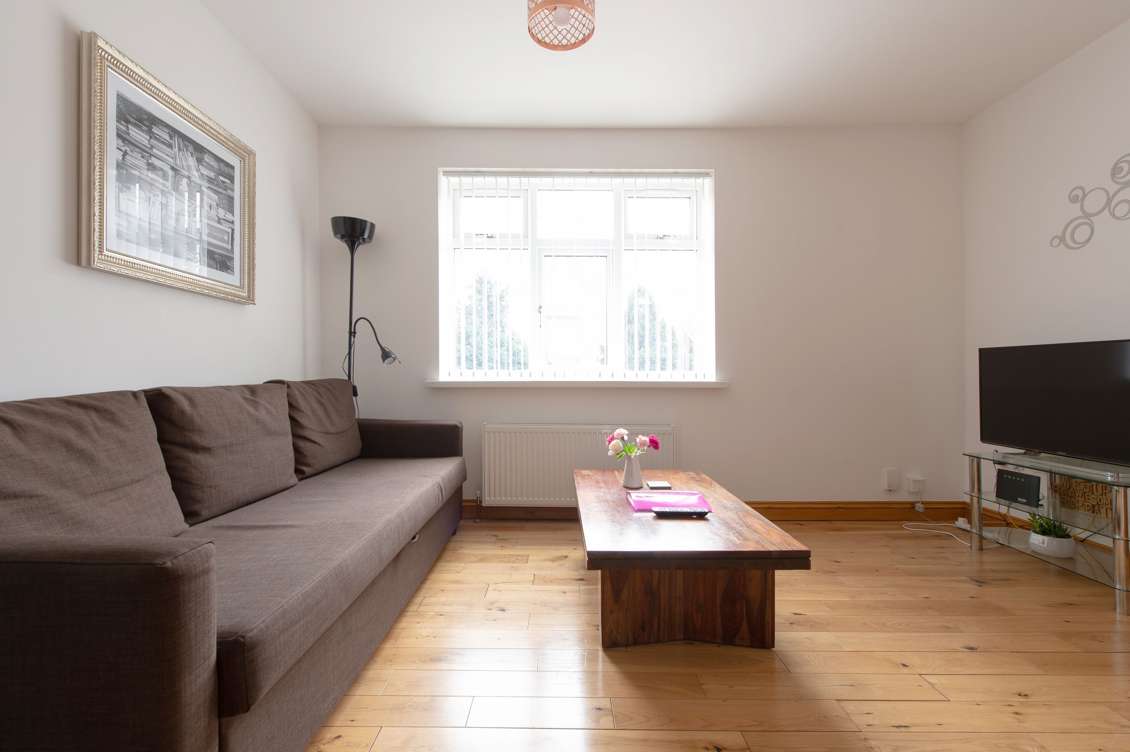 A living room with a brown sofa, a wooden coffee table with a vase of flowers, a large window, and a flat-screen TV on a glass stand.